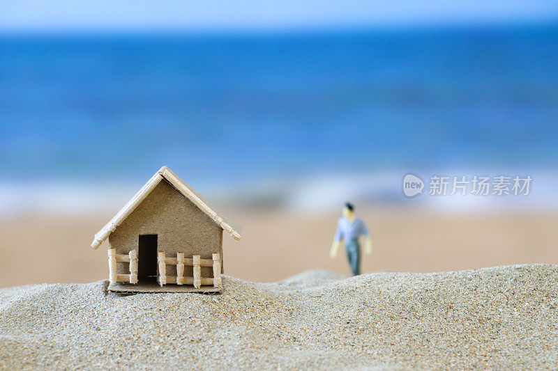 特写图像的微型，男性雕像除了纸板模型度假小屋在一堆沙子，沙滩背景与海浪在低潮，大海和海岸线，重点在前景，度假租赁和度假屋的概念，复制空间