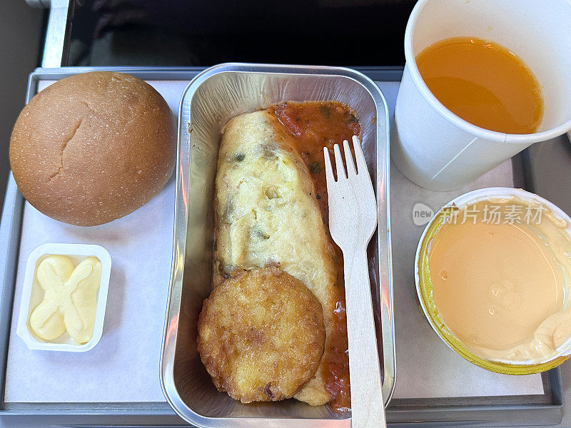 飞机食品托盘的全画幅图像，里面有早餐，锡纸托盘里的煎蛋卷，西红柿和洋葱酱和土豆泥，白面包卷和黄油部分，一罐酸奶，塑料杯橙汁，木制叉子，高架视图