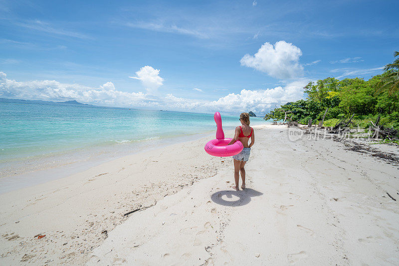一名年轻女子与充气火烈鸟在泰国群岛田园诗般的海滩上散步。人们旅游目的地有趣和酷的态度概念
