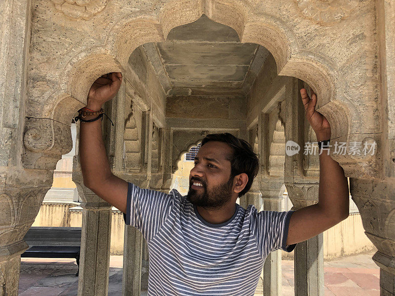 年轻英俊的印度男子在印度观光旅游手举过头顶，在有柱子的寺庙的石拱上休息，亚洲印度教印度男子与破洞牛仔t恤摆姿势，在德里阴凉炎热的天气休息