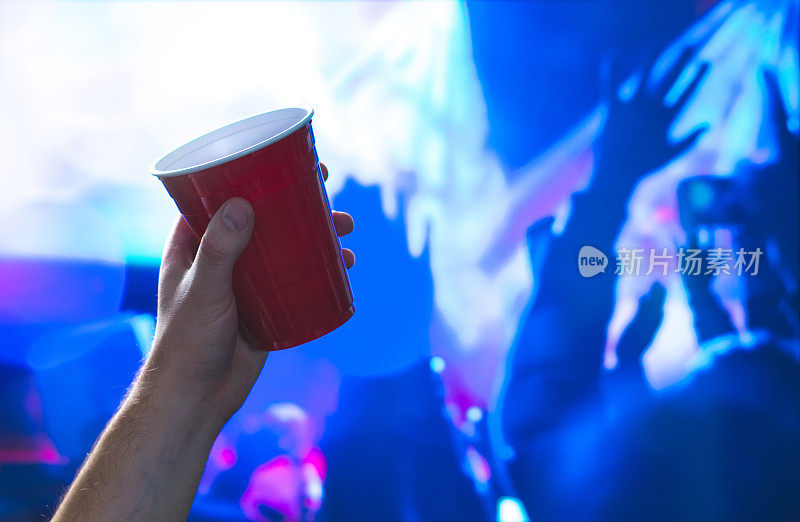 一个年轻人在夜总会的舞池里拿着红色的派对杯。在迪斯科舞厅，手里拿着酒精容器。大学生在玩，在跳舞。在后面庆祝的人们。活动营销和推广。