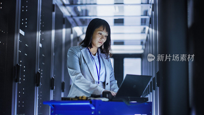 亚洲女性IT工程师工作在一个笔记本电脑上的工具推车，她扫描硬盘。她在一个满是机架服务器的大数据中心。