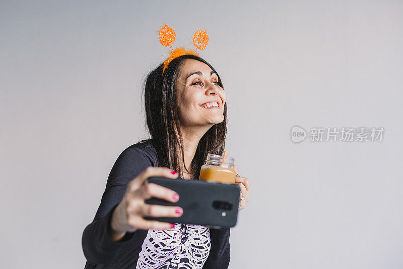 年轻漂亮的女人喝着橙汁，用手机自拍。穿着黑白骷髅服装。万圣节的概念。在室内