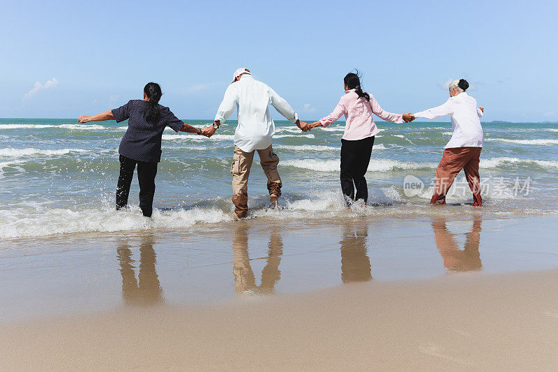 四名乐天派老前辈朋友一起外出旅游，手牵着手站在局部海滩上，尽情玩乐，享受时光，在假日里休息放松。