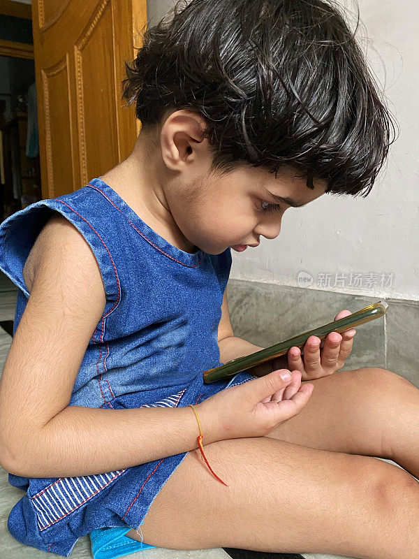 图为年轻的印度女孩坐在地板上专注地盯着手机屏幕，聚精会神地盯着智能手机
