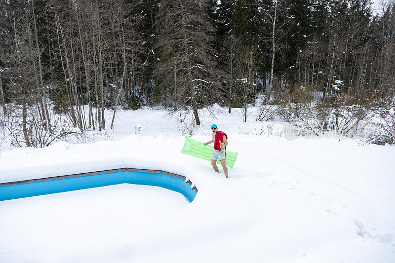 一个年轻人站在泳裤和气垫在他的手。一名男子站在积雪覆盖的室外游泳池旁。