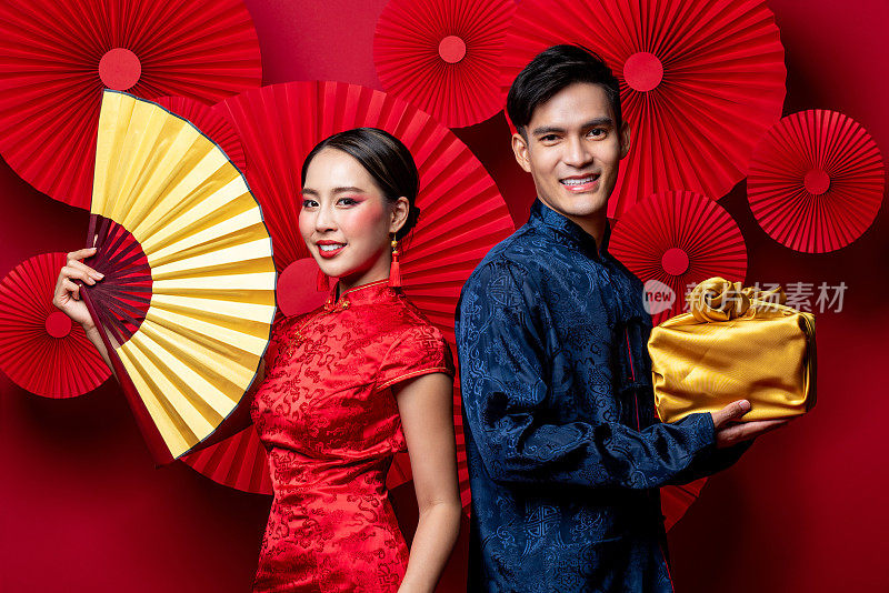身着传统服饰的亚洲夫妇面带微笑，以东方红色为背景装饰，以扇子和金色包装作为春节礼物