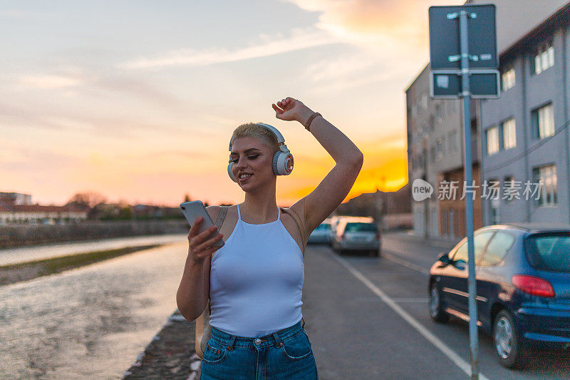 美丽的年轻女子与短金发享受音乐在她的耳机，而步行通过一个城市在日落