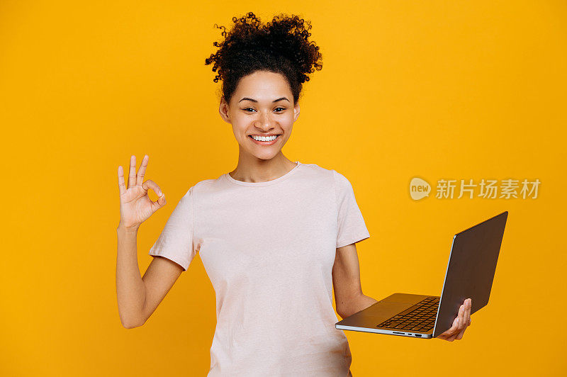 快乐漂亮，卷发的非洲裔美国年轻女性，穿着休闲的衣服，手里拿着一台打开的笔记本电脑，看着镜头，做出OKAY的手势，同意的手势，站在孤立的橙色背景上