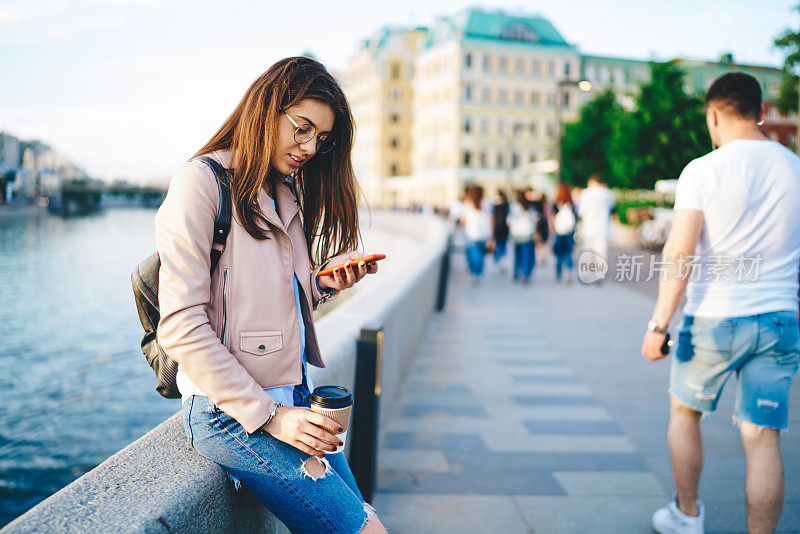 年轻女子在旅行中使用4G连接在手机上聊天，站在江城大桥上用智能手机使用导航和地图