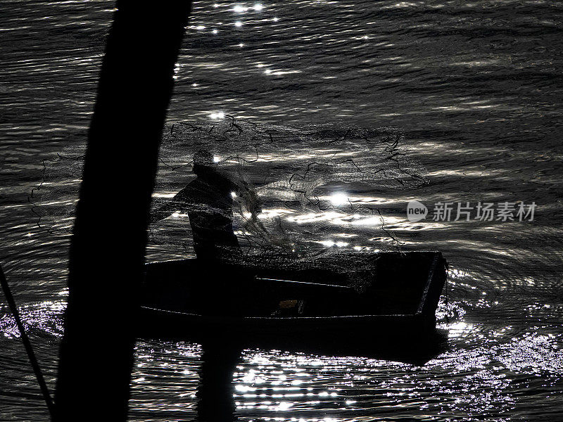 一个渔夫站在他的船上，向水中撒网的剪影。