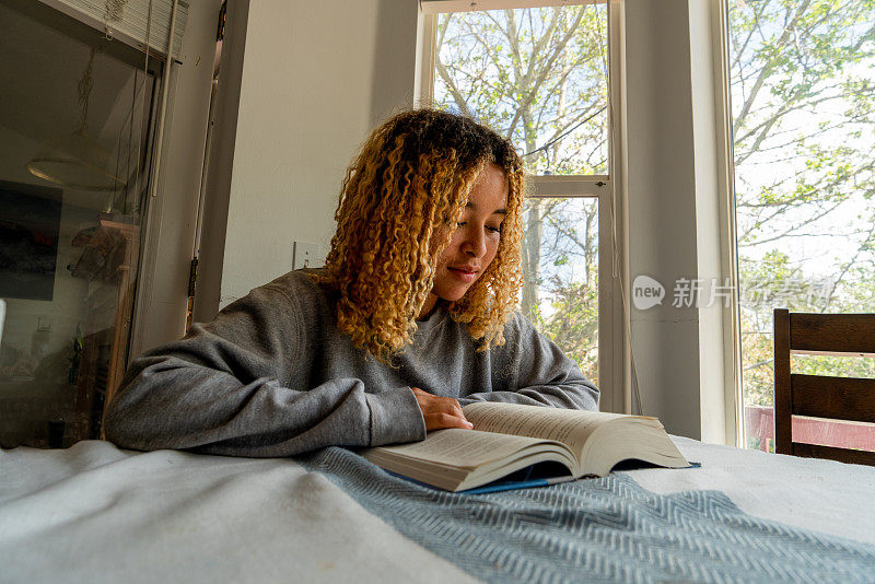 一个十几岁的非裔美国女孩，一头齐肩长发，全神贯注地看书，她的思想被书页上的文字所吸引，她舒适地穿着一件灰色运动衫，坐在餐桌旁。