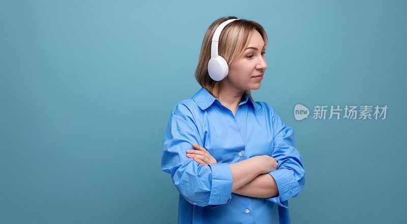 宽屏照片上，一个沉思的欧洲女孩穿着休闲衬衫，戴着白色的大耳机，在一个蓝色的孤立背景上听音乐