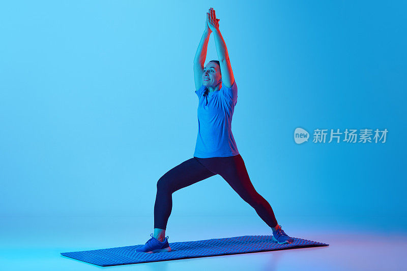 保持身体健美。女人在健身matt上训练，在霓虹灯的蓝色工作室背景下伸展身体