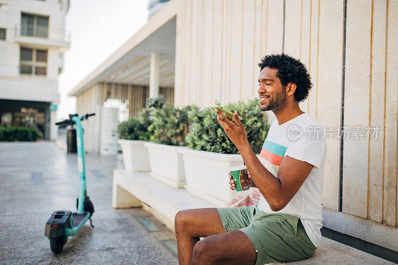 一个黑人坐在长凳上，用一次性杯子喝咖啡，休息一下，骑着电动滑板车