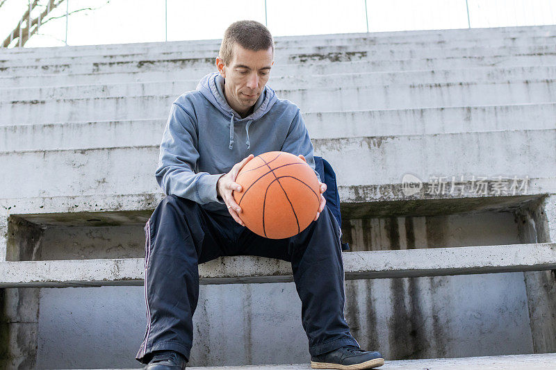 一名男子拿着篮球坐在篮球场旁边的台阶上，赛后休息。