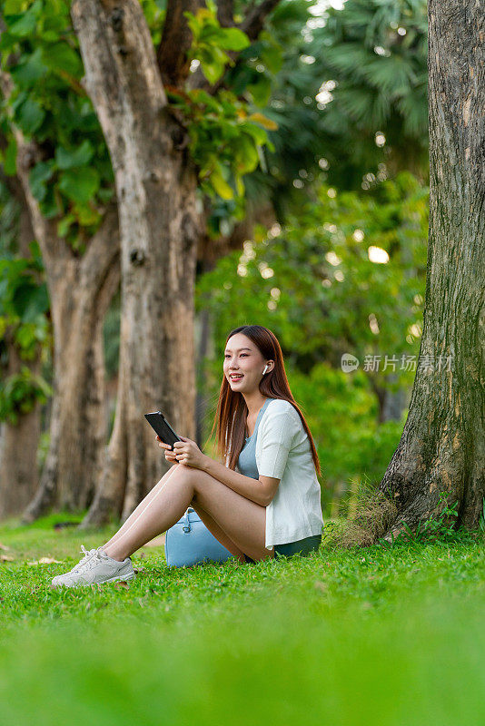 Z世代亚洲女性在公园树下使用耳机和平板电脑