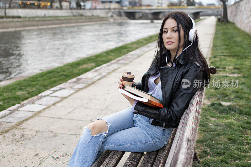 年轻漂亮的女大学生背着背包在河边玩。她戴着耳机，穿着皮夹克，等待着大学课程的开始。