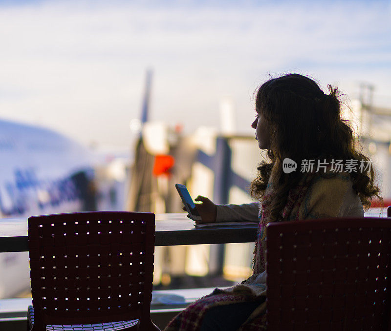 一名少女在机场的咖啡馆里摆弄智能手机，背景是一架飞机