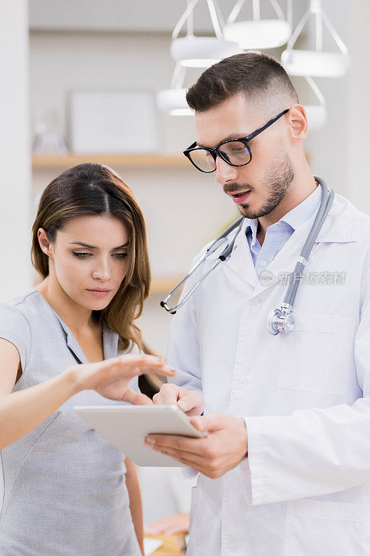 严肃专注的女性诊所经理一边和年轻医生聊天，一边解释如何在平板电脑上使用手机应用程序