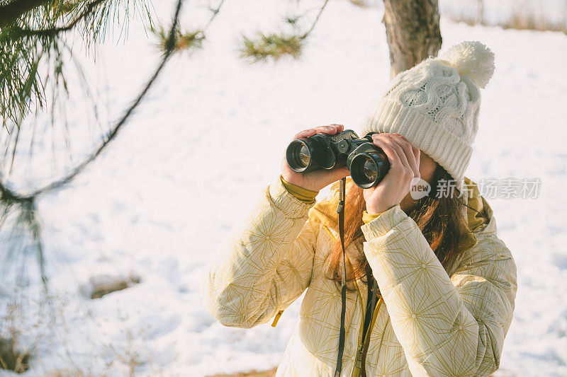 冬天的森林里，戴着白帽、背着背包的女孩用双筒望远镜看东西