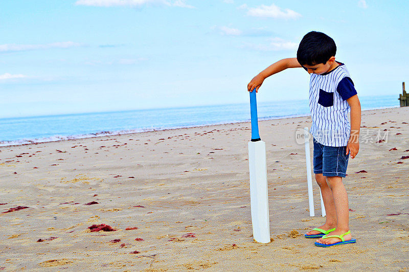 水平的照片，一个来自印度的小男孩站在海滩上，拿着板球棒守卫，在一个炎热的夏天。