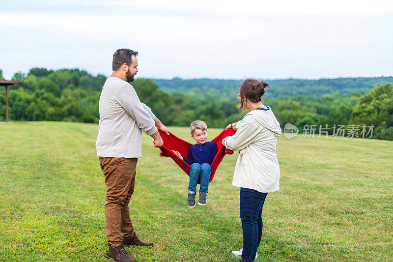 年轻夫妇和儿子玩作为一个家庭在公共草地绿色公园设置一起玩