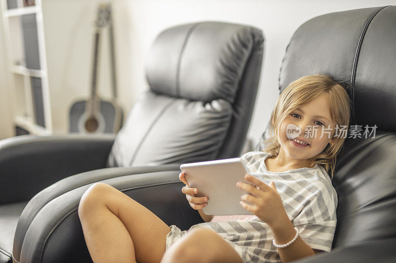 微笑的小女孩穿着睡衣，坐在扶手椅上，在家里使用数码平板电脑