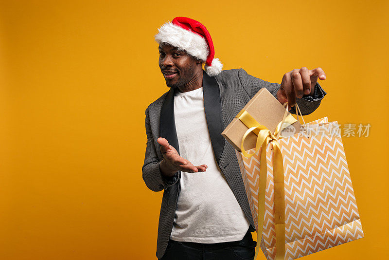 一个戴着圣诞帽、皮肤黝黑的男人微笑着，对着镜头拿出一个礼物袋