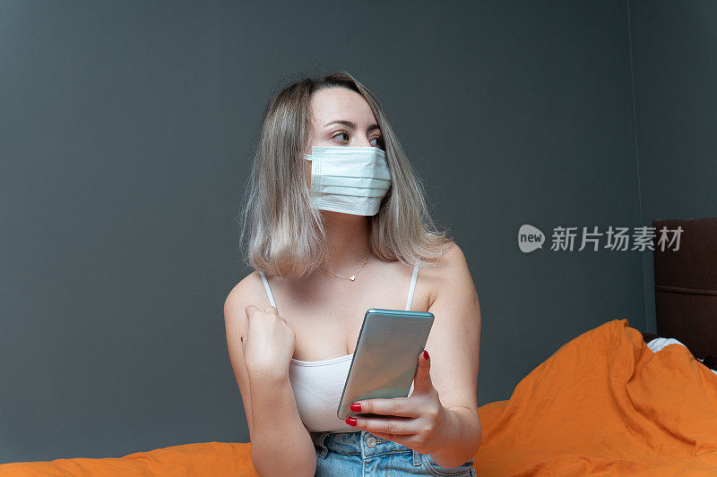 年轻美丽的女人用智能手机戴着外科口罩在床上的肖像