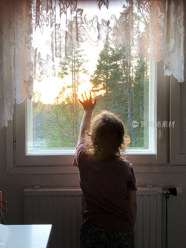 一个5岁的小女孩站在窗前看日落。从后面可以看到这个孩子。孩子用他的手抓住了太阳和梦想。