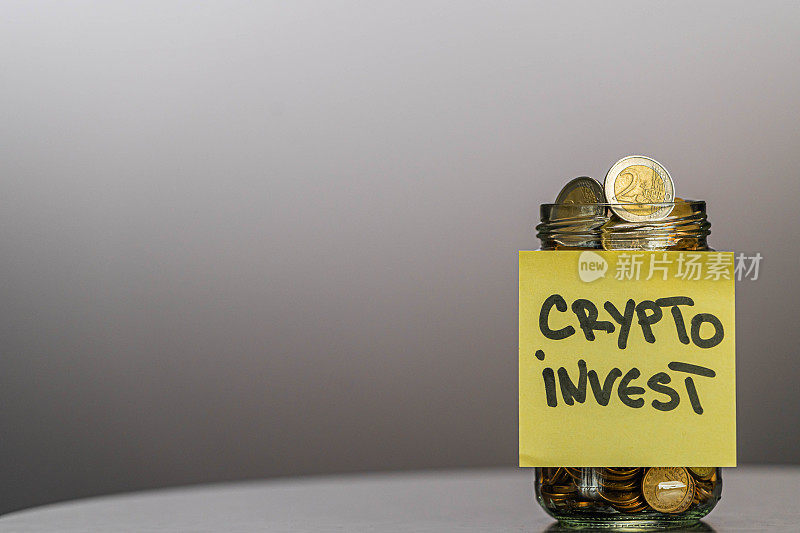 一个装满硬币和两枚欧元硬币的玻璃罐子，上面有加密货币投资文字，灰色背景