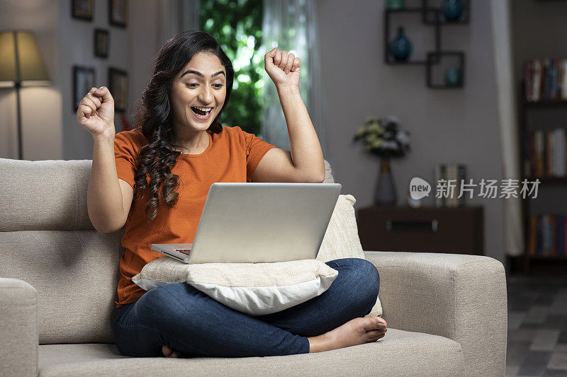 兴奋的年轻女子举起拳头坐在沙发上，同时使用笔记本电脑在家里:库存照片