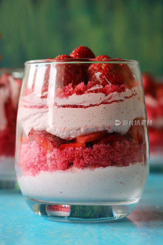 这是一幅单独的玻璃特写，里面有自制的草莓慕斯甜点，上面覆盖着整个草莓，奶油慕斯的分层布丁和红丝绒蛋糕屑，蓝绿色的表面和斑纹的绿色背景