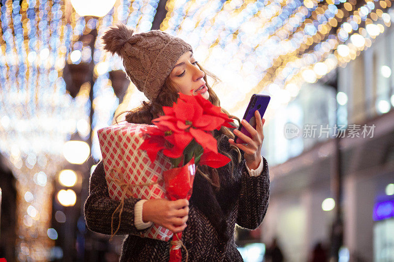 美丽的女人使用电话和持有圣诞礼物户外