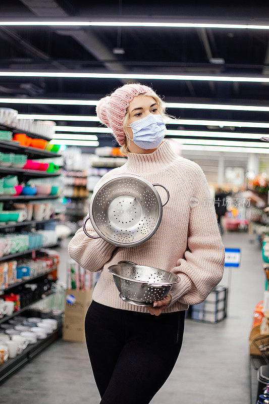 漂亮的年轻女子戴着面具，戴着粉红色的针织帽和毛衣，在超市的厨房用具部和两个滤器摆出幽默的姿势。购物在大流行。积极的情感。幽默。