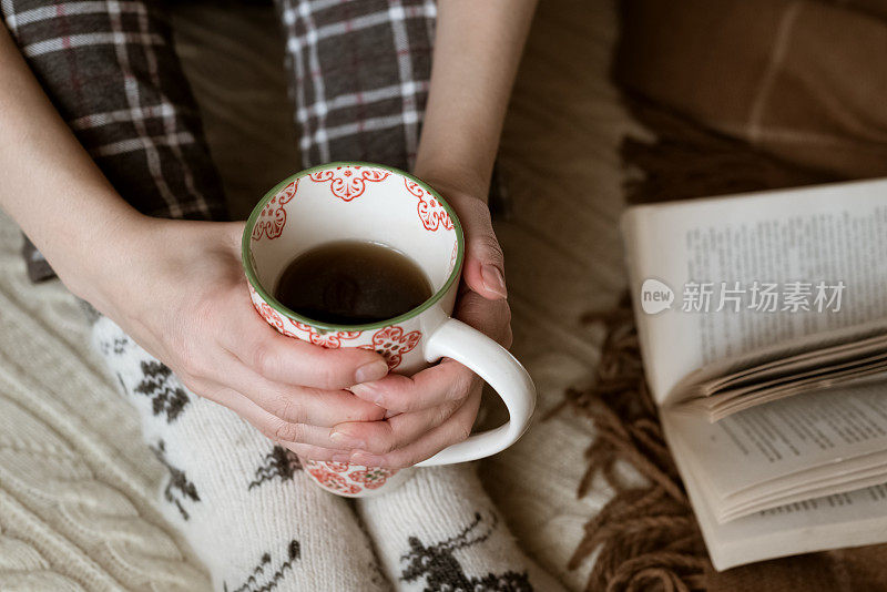 女人穿着暖和的羊毛针织袜子，坐在沙发上，旁边是一本打开的书，手里拿着一杯茶