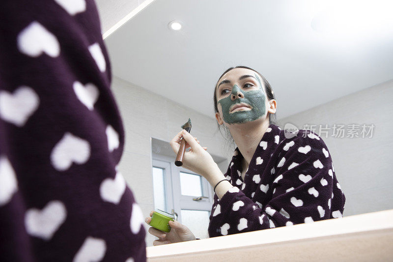 一名年轻女子用刷子在家用浴室的镜子前敷绿色面膜，从低角度看