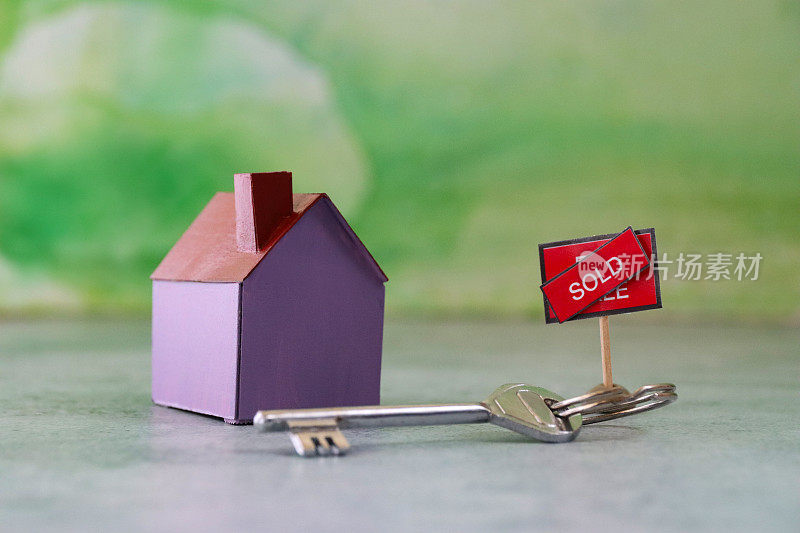 钥匙圈上的钥匙旁边的紫色纸板房子的形象，“出售”的标志与对角线旁边的“出售”横过文字横幅，斑驳的绿色背景，房地产和搬家的概念