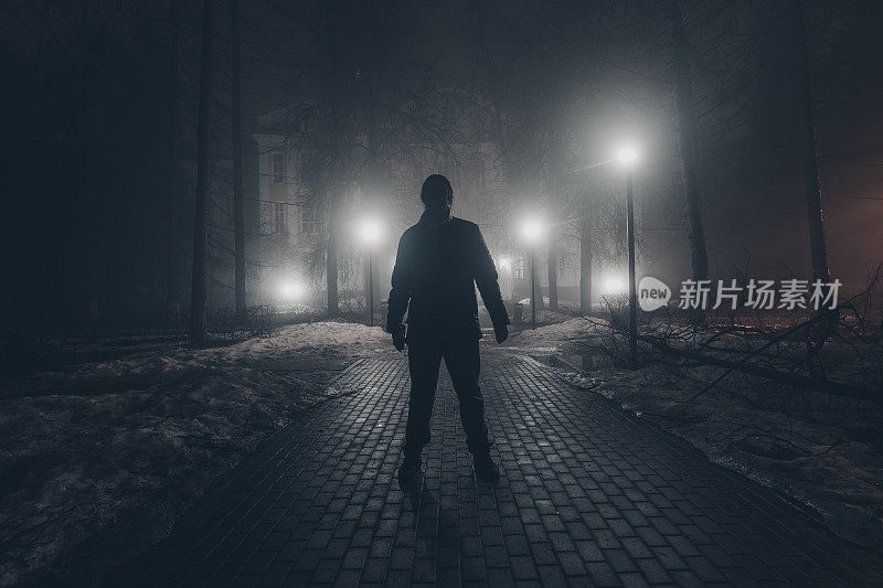 悲伤的人独自走在夜晚雾蒙蒙的公园里的小巷里。后视图