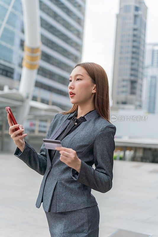 一名年轻的亚洲女商人穿着灰色的办公套装，在商业区用她的智能手机使用信用卡进行在线交易，她微笑着自信