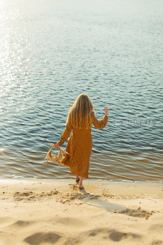 后面的观点。穿黄色衣服的金发女人躺在河岸上。夏天的一天。长头发的。沙子。