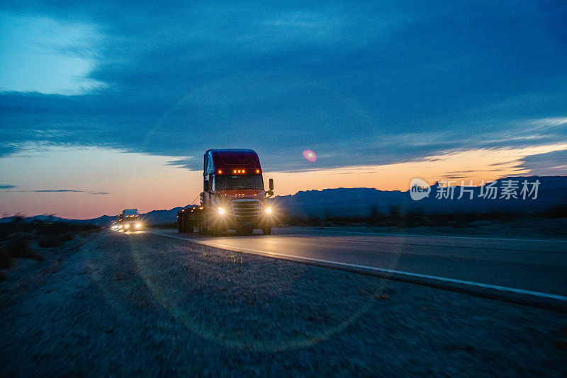日落时分，一辆18轮大卡车隆隆驶过内华达州的沙漠，扬起尘土。群山巍然矗立在背景中，当卡车在渐暗的光线中行驶时，它们投下了长长的影子。