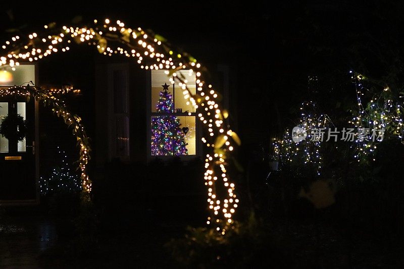 花园的拱门上有灯光，背景是一棵挂在房子窗户上的圣诞树