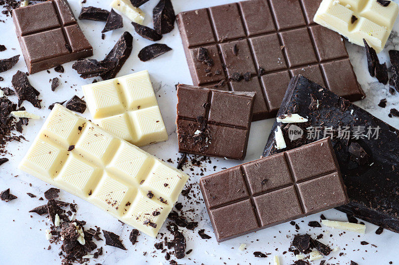 全帧图像的巧克力棒，牛奶巧克力块，白巧克力方块和黑巧克力块与磨碎的刨花在白色大理石效果的背景，抬高视图