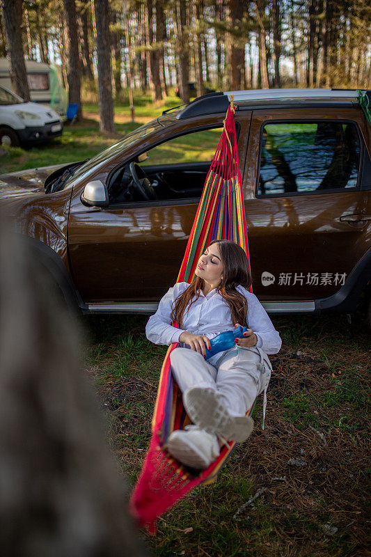 在大自然中享受的年轻女子，她躺在吊床上，拿着一瓶水在露营时，背景是露营拖车和越野车