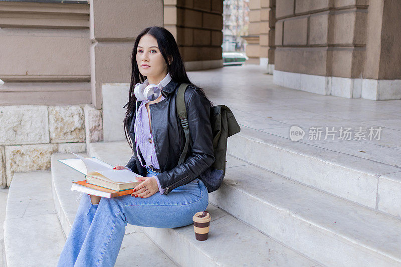 年轻漂亮的女大学生背着双肩包坐在大学楼梯上。她戴着耳机，穿着皮夹克，等待着大学课程的开始。