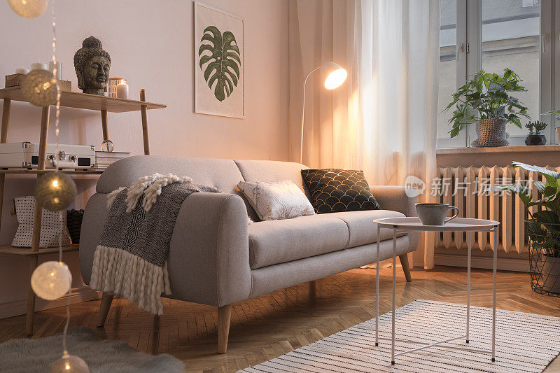 时尚的斯堪的纳维亚室内设计沙发，海报，植物，枕头，书架，咖啡桌，舒适的毯子和模拟框架。白色背景墙，棕色镶木地板。晚上的照片。