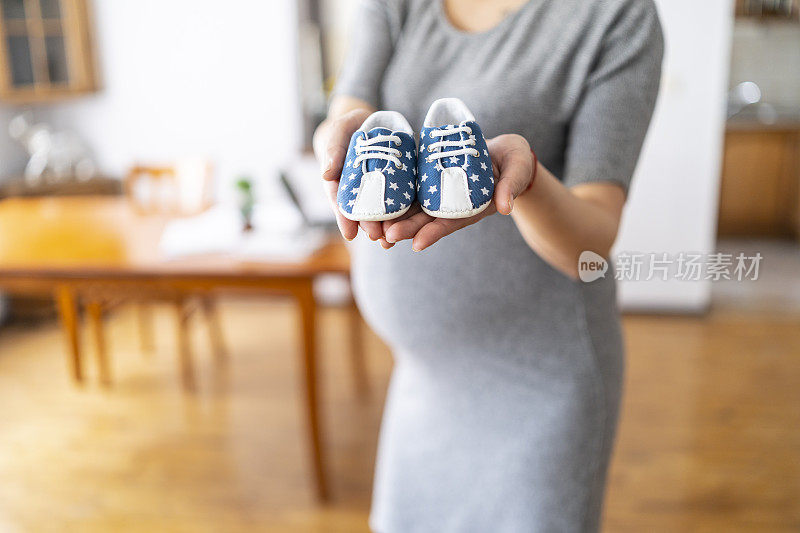 孕妇站着，手里拿着蓝色婴儿鞋