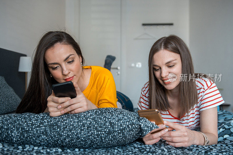 两个朋友躺在床上一边聊天一边玩智能手机